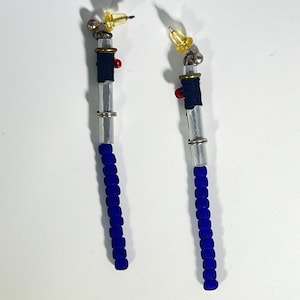 Puede incluir: Un par de pendientes hechos a mano con un dise&ntilde;o &uacute;nico. Cada pendiente presenta una base de cuentas azules, una secci&oacute;n central transparente y una secci&oacute;n envuelta en azul marino. Los pendientes tienen un acento rojo y una parte superior dorada con un poste amarillo.