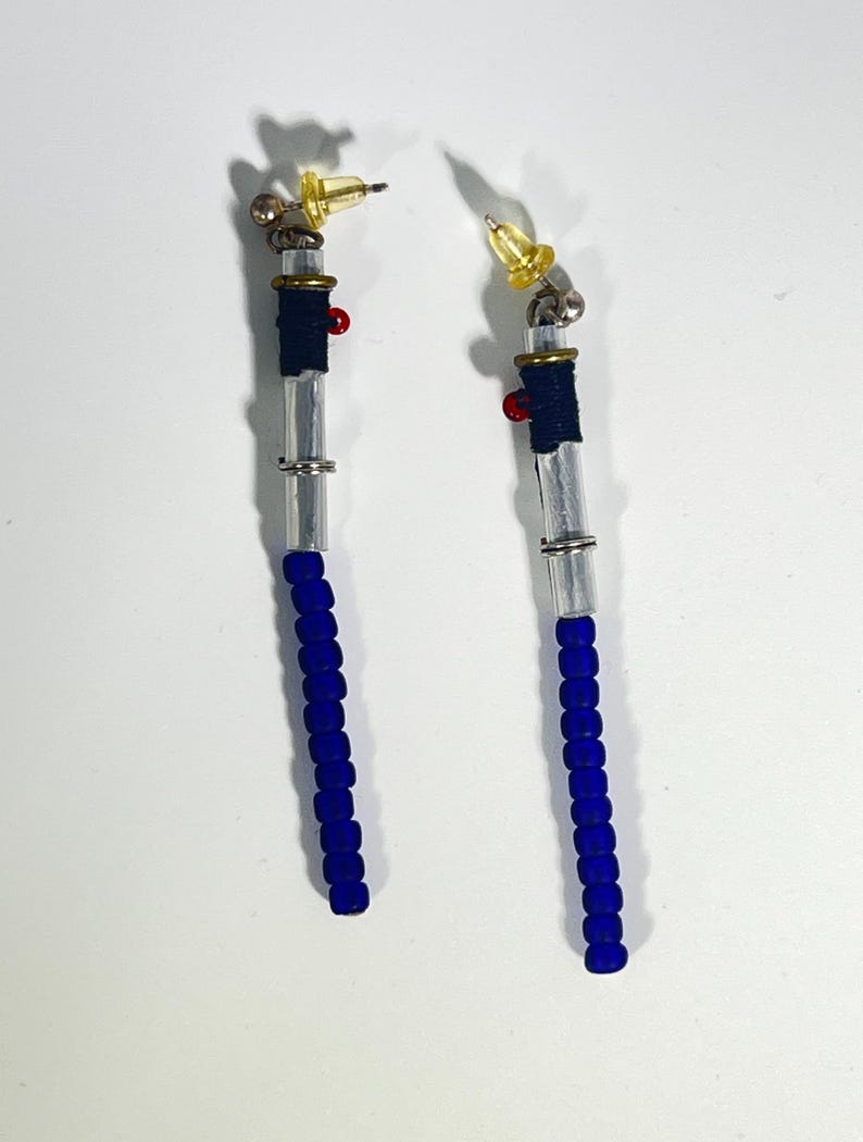 Puede incluir: Un par de pendientes hechos a mano con un dise&ntilde;o &uacute;nico. Cada pendiente presenta una base de cuentas azules, una secci&oacute;n central transparente y una secci&oacute;n envuelta en azul marino. Los pendientes tienen un acento rojo y una parte superior dorada con un poste amarillo.