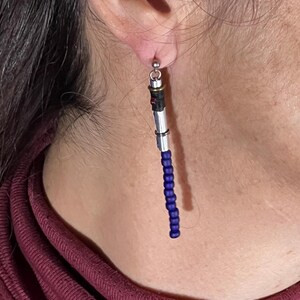 Puede incluir: Un solo pendiente con un dise&ntilde;o de sable de luz. La parte superior es plateada con detalles en negro y rojo. La secci&oacute;n media es plateada, y la parte inferior es una hebra de cuentas azul oscuro. El pendiente cuelga de un poste plateado.