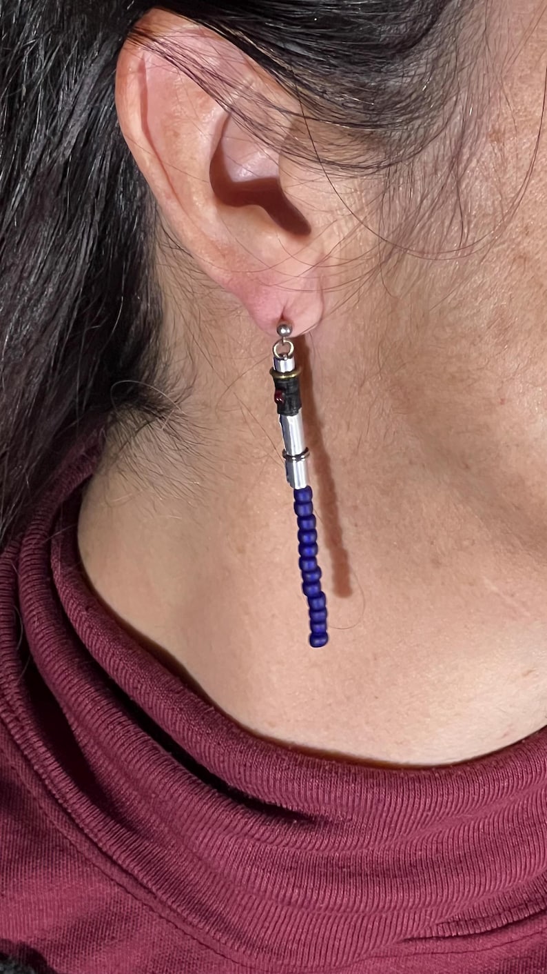 Puede incluir: Un solo pendiente con un dise&ntilde;o de sable de luz. La parte superior es plateada con detalles en negro y rojo. La secci&oacute;n media es plateada, y la parte inferior es una hebra de cuentas azul oscuro. El pendiente cuelga de un poste plateado.