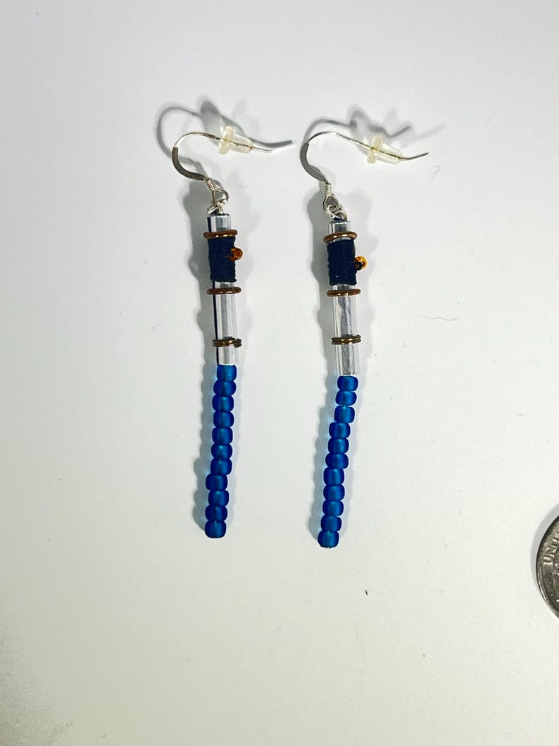 Puede incluir: Un par de pendientes hechos a mano. Cada pendiente presenta un gancho plateado, un tubo transparente y una banda azul oscuro. La parte inferior est&aacute; adornada con peque&ntilde;as cuentas azules vibrantes. Los pendientes est&aacute;n dise&ntilde;ados para colgar.