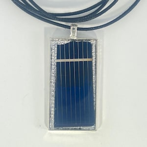 Puede incluir: Un colgante rectangular con un diseño de panel solar azul oscuro, enmarcado en plata con un borde de cuentas, cuelga de un collar de cordón negro. El diseño del colgante presenta líneas verticales y una argolla plateada.