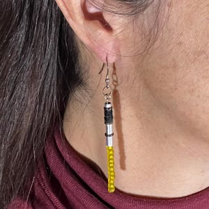 Puede incluir: Pendientes colgantes con dise&ntilde;o de sable de luz. Los pendientes presentan un mango de metal plateado, detalles negros y plateados y una hoja de cuentas amarillas. Cuelgan de un gancho plateado.