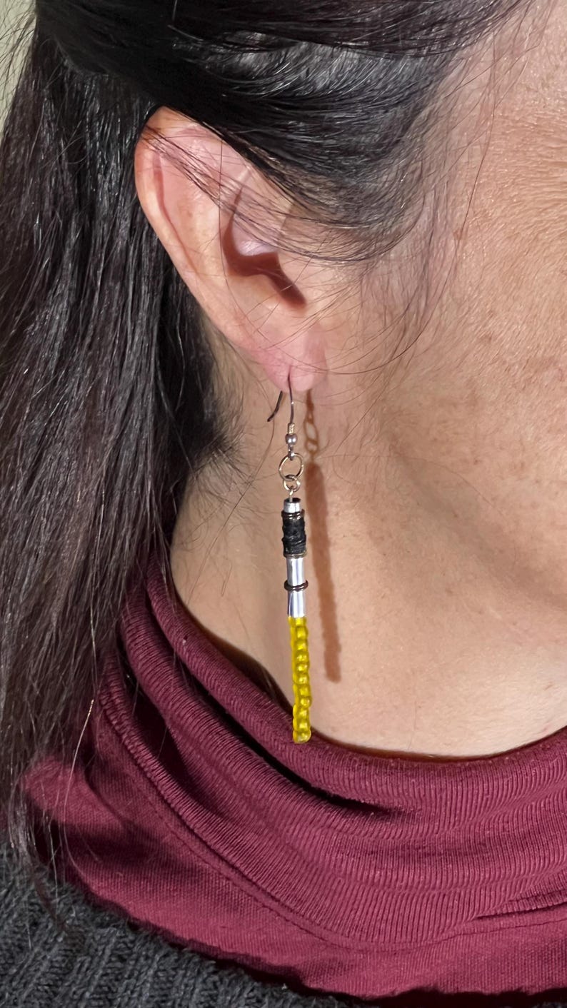 Puede incluir: Pendientes colgantes con dise&ntilde;o de sable de luz. Los pendientes presentan un mango de metal plateado, detalles negros y plateados y una hoja de cuentas amarillas. Cuelgan de un gancho plateado.