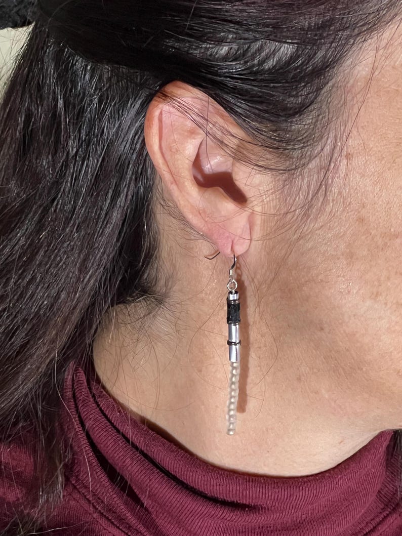 Puede incluir: Pendientes colgantes con cuentas plateadas y negras. Los pendientes tienen un gancho plateado, una secci&oacute;n cil&iacute;ndrica negra y plateada y una larga hebra de peque&ntilde;as cuentas de color claro. Los pendientes cuelgan de una oreja.