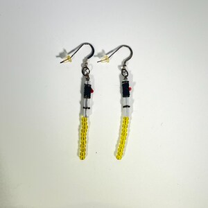 Puede incluir: Un par de pendientes hechos a mano con un dise&ntilde;o de sable de luz. Los pendientes tienen cuentas amarillas, transparentes, negras y rojas. Los pendientes tienen un gancho de color dorado.