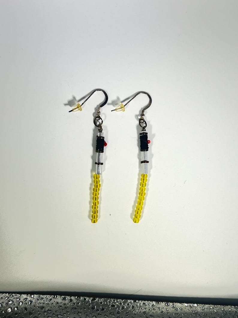 Puede incluir: Un par de pendientes hechos a mano con un dise&ntilde;o de sable de luz. Los pendientes tienen cuentas amarillas, transparentes, negras y rojas. Los pendientes tienen un gancho de color dorado.