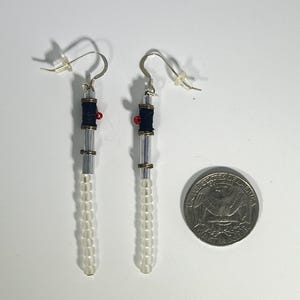 Puede incluir: Un par de pendientes hechos a mano con un dise&ntilde;o de sable de luz. Los pendientes presentan cuentas cil&iacute;ndricas transparentes, hilo negro y detalles rojos. Una moneda est&aacute; al lado para la comparaci&oacute;n de tama&ntilde;o.