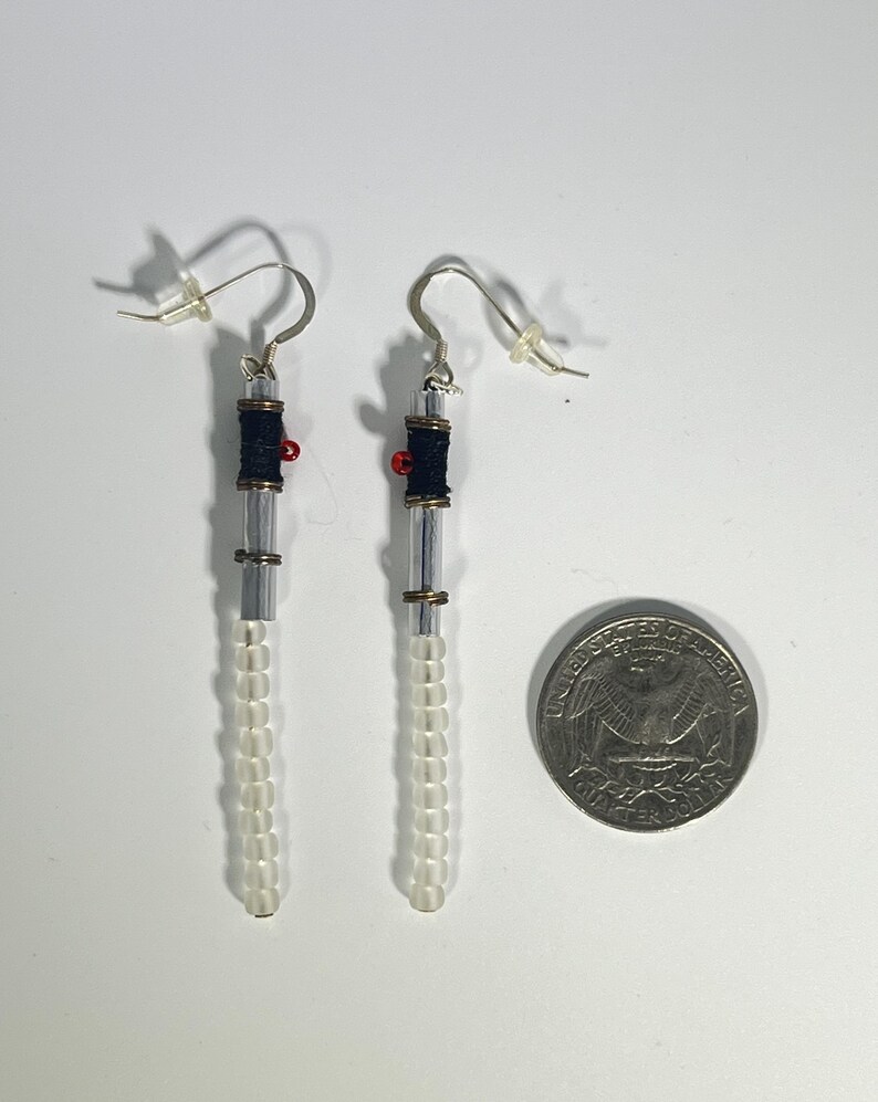 Puede incluir: Un par de pendientes hechos a mano con un dise&ntilde;o de sable de luz. Los pendientes presentan cuentas cil&iacute;ndricas transparentes, hilo negro y detalles rojos. Una moneda est&aacute; al lado para la comparaci&oacute;n de tama&ntilde;o.