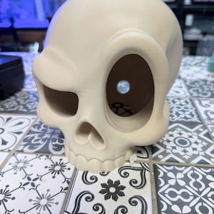 May include: A cream-coloured, stylised skull sculpture with exaggerated, asymmetrical eye sockets. The skull features a smooth, matte finish and a hollow interior, with a small black symbol visible on the inner base of the cranium through the large left eye socket.