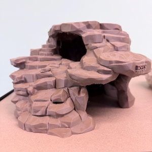 May include: A brown miniature cave structure with a textured, rocky exterior. The cave has an arched entrance and a small staircase leading up to a higher level. The overall design is detailed and realistic.