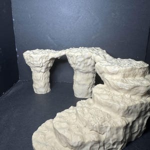 May include: Off-white miniature terrain pieces, resembling rock formations and a cave arch. The pieces have a textured surface and are arranged on a dark surface. The scene includes a cave arch and a rock formation.