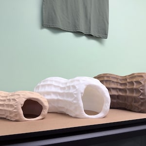 May include: Three hollow, peanut-shaped ceramic sculptures arranged in a row. The leftmost sculpture is light tan, the middle one is white, and the rightmost one is brown. Each piece features a textured, dimpled surface and a large open end.