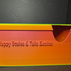 May include: A rectangular, multi-colored storage box with a curved opening. The box is primarily orange with a pink interior and a green and yellow trim. The text "Happy Scales & Tails Exotics" is embossed on the front.