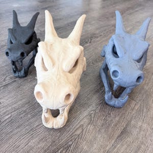 May include: Three dragon skull sculptures arranged in a group. One skull is matte black, one is a light cream colour, and one is a muted blue-grey. Each skull features prominent horns, hollow eye sockets, a wide snout, and sharp teeth in the open jaw.
