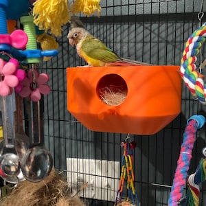 May include: A small green and yellow bird perched on top of a bright orange plastic nesting box with a circular opening. The box is mounted inside a wire cage filled with colourful hanging toys, including plastic flowers and a twisted rope.