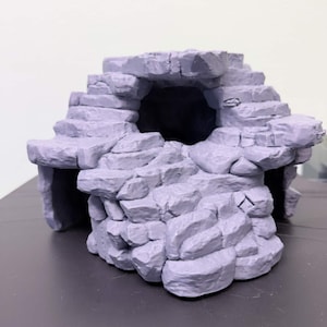 Könnte beinhalten: Eine graue Miniatur-Höhlenstruktur mit einer zentralen Öffnung und geschichteten Felsformationen. Die Höhle hat eine strukturierte Oberfläche und scheint aus Stein zu bestehen. Das Design umfasst Stufen und einen abgerundeten Bogen, geeignet für Tabletop-Spiele oder Ausstellungen.
