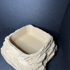 May include: A beige reptile feeding dish, designed to resemble a rock. It features a square opening and a textured exterior. The interior is smooth, and the design is intended for a reptile habitat, with measurements in cm.