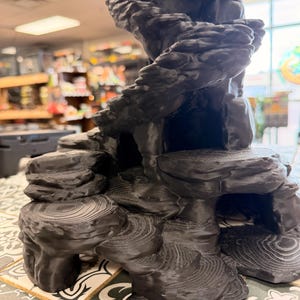 May include: A black, three-dimensional printed structure featuring a multi-tiered, rocky landscape design. The object displays layered, circular textures resembling tree rings on its flat surfaces and includes several hollow cave-like openings.