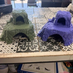 May include: Two small, textured reptile hides shaped like rocky caves with arched openings. One hide is matte olive green, and the other is matte purple. Both structures feature a tiered design with a flat upper platform and a hollow interior.