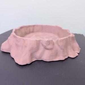 May include: A light pink, low-profile, circular dish with a textured, uneven exterior surface that mimics the appearance of natural rock formations. The object features a wide, shallow basin with a smooth, slightly rippled interior base.