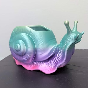 Pode incluir: Um vaso de plantas em forma de caracol com cores gradientes. A concha transita de verde-azulado para roxo e rosa. O corpo do caracol é verde-azulado com detalhes texturizados. Um item decorativo único.