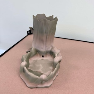 May include: A 3D printed desk organizer shaped like a tree stump with a hollow cylindrical top and a wide, shallow basin at the base. The object features a gradient color scheme transitioning from muted pink to sage green, with a smooth texture.