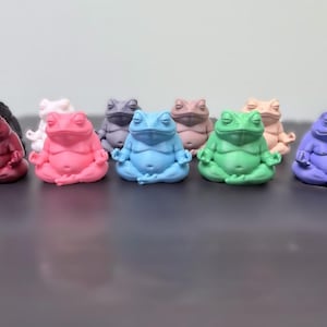 May include: A collection of nine small frog figurines in various solid colors including red, pink, blue, green, purple, black, white, grey, and tan. Each frog is depicted in a seated meditation pose with eyes closed and hands resting on knees.