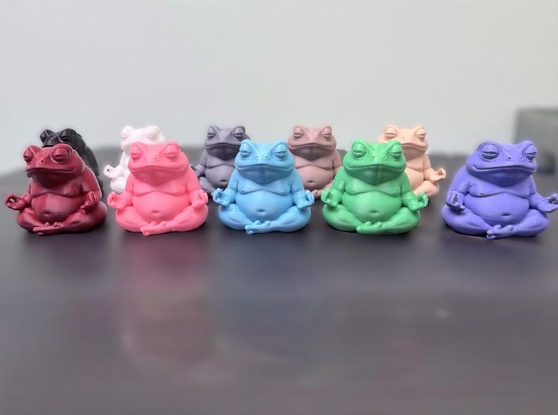 May include: A collection of nine small frog figurines in various solid colors including red, pink, blue, green, purple, black, white, grey, and tan. Each frog is depicted in a seated meditation pose with eyes closed and hands resting on knees.