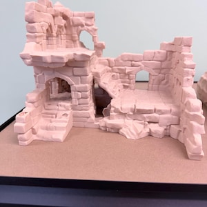 May include: A light pink miniature castle ruin with arched doorways, stairways, and weathered brickwork. The model has a detailed, textured surface, suggesting an aged stone structure. The castle is designed for tabletop gaming or model building.
