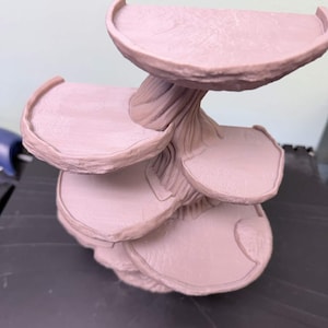 May include: A light pink, tiered display stand with a tree-like trunk and several rounded platforms. The platforms have a curved edge. The stand is made of a solid material and is suitable for displaying small items.