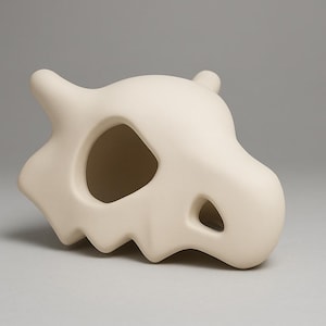 May include: A matte off-white ceramic sculpture shaped like a stylised animal skull. The object features a smooth, rounded surface with a large, irregular eye socket opening and a smaller triangular nasal cavity, with two small horn-like protrusions.