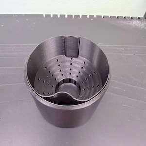 May include: A matte black, cylindrical container featuring a removable inner basket with a perforated base and sides. The inner component has a distinct curved cutout along the top rim, while the outer shell maintains a smooth, solid wall design.