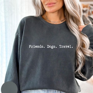 May include: Dark gray sweatshirt with the text "Friends. Dogs. Travel." in white. The sweatshirt has a crew neck and long sleeves. The image shows a person wearing the sweatshirt.