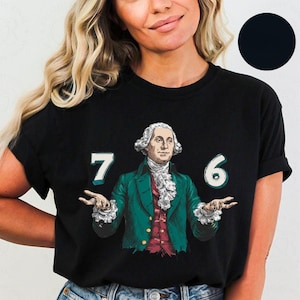 Op de afbeelding: Zwart T-shirt met een afbeelding van George Washington in een groene jas en rood vest, met de cijfers "7" en "6". Het ontwerp heeft een vintage, geïllustreerde stijl.