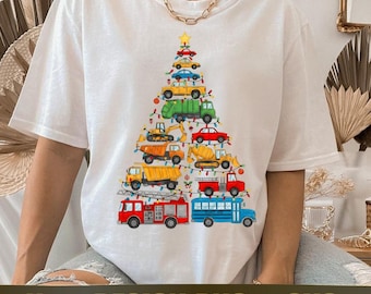Op de afbeelding: Wit T-shirt met een kerstboomontwerp gemaakt van verschillende kleurrijke voertuigen, waaronder een brandweerwagen, schoolbus en dumper. Het ontwerp bevat een ster en lichtslingers. De tekst onderaan luidt "New DESIGN PNG & SVG".