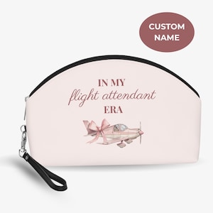 Peut inclure: Une trousse de maquillage rose clair avec une fermeture éclair et une poignée noires. Le sac porte l'inscription "IN MY flight attendant ERA" et une illustration à l'aquarelle d'un petit avion avec un nœud.