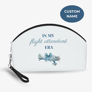 Könnte beinhalten: Weiße Kosmetiktasche mit schwarzem Reißverschluss und Handschlaufe. Die Tasche trägt den Text "IN MY flight attendant ERA" in blauer Schrift sowie eine Aquarellillustration eines Oldtimer-Flugzeugs mit blauer Schleife. Ein blauer Kreis mit "CUSTOM NAME" befindet sich oben rechts.