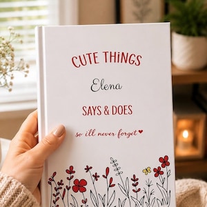 Op de afbeelding: Een wit hardcover dagboek met rode en zwarte tekst die luidt CUTE THINGS Elena SAYS & DOES so ill never forget. De onderkant van de kaft bevat een kleurrijke illustratie van rode en gele bloemen met zwarte stengels en bladeren.