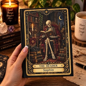 May include: A black hardcover book titled The Reader with the name Sophia printed on the cover. The cover art features a skeleton sitting in a chair reading a book in a library setting with a black cat, gold filigree borders, and celestial motifs.