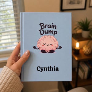 May include: A light blue hardcover notebook featuring the text "Brain Dump" above a cartoon illustration of a pink brain character sitting in a meditative pose. The name "Cynthia" is printed in black text at the bottom of the front cover.