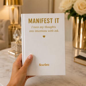 May include: A white hardcover journal with gold foil text that reads MANIFEST IT, I turn my thoughts into intentions with ink., a small gold heart icon, and the name Scarlett printed at the bottom. The cover has a clean, smooth finish.