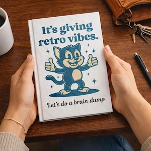 Könnte beinhalten: Ein offenes weißes Notizbuch mit dem Text "It's giving retro vibes." und "Let's do a brain dump". Das Cover zeigt eine Cartoon-Katze, die einen Daumen hoch gibt. Eine Kaffeetasse, ein Stift und Haftnotizen liegen auf der Holzoberfläche.