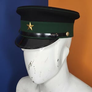 May include: A black and green military-style hat with a gold star emblem. The hat features a shiny black visor and a green band. A gold button is visible on the side. The hat is displayed on a white mannequin head against a blue and orange background.