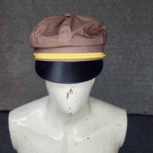 May include: A brown, structured cap with a black brim and a gold band. The cap has a rounded crown and a flat, stiff brim. The gold band is positioned just above the brim, adding a decorative element. The cap is displayed on a mannequin.
