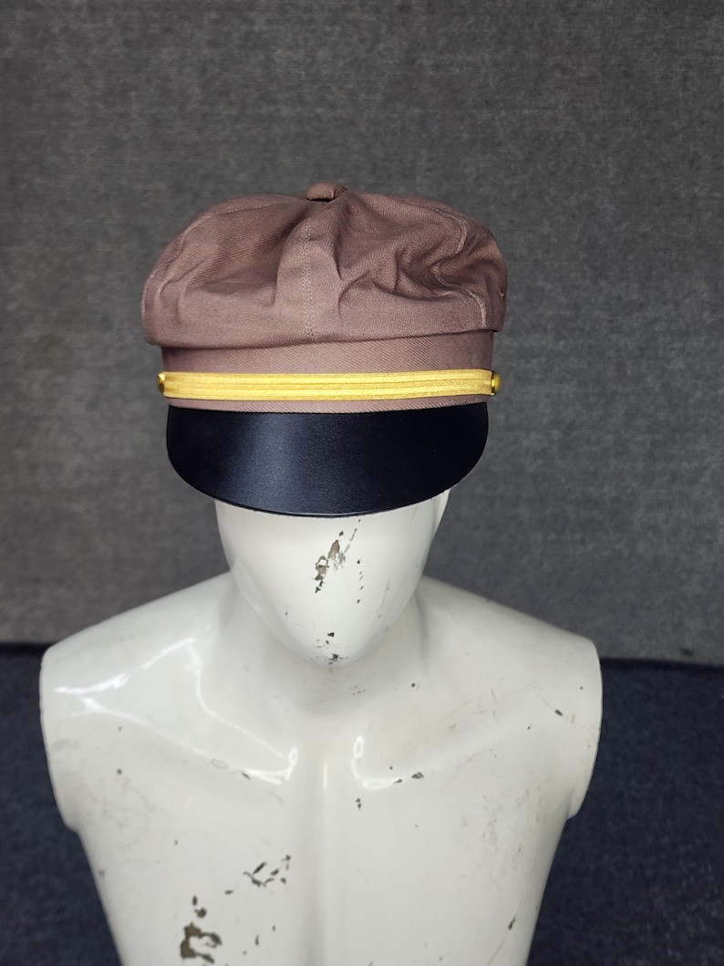 May include: A brown, structured cap with a black brim and a gold band. The cap has a rounded crown and a flat, stiff brim. The gold band is positioned just above the brim, adding a decorative element. The cap is displayed on a mannequin.
