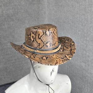 May include: A cowboy hat featuring a tan and black snakeskin pattern. The hat includes a decorative band with a braided tan centre and black edges, along with a thin black chin strap that hangs down from the brim of the hat.