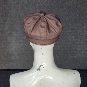 May include: A brown newsboy cap with a rounded crown and a small brim. The cap is made of a soft, textured fabric and has a button detail at the top. The cap is displayed on a white mannequin head against a gray background.