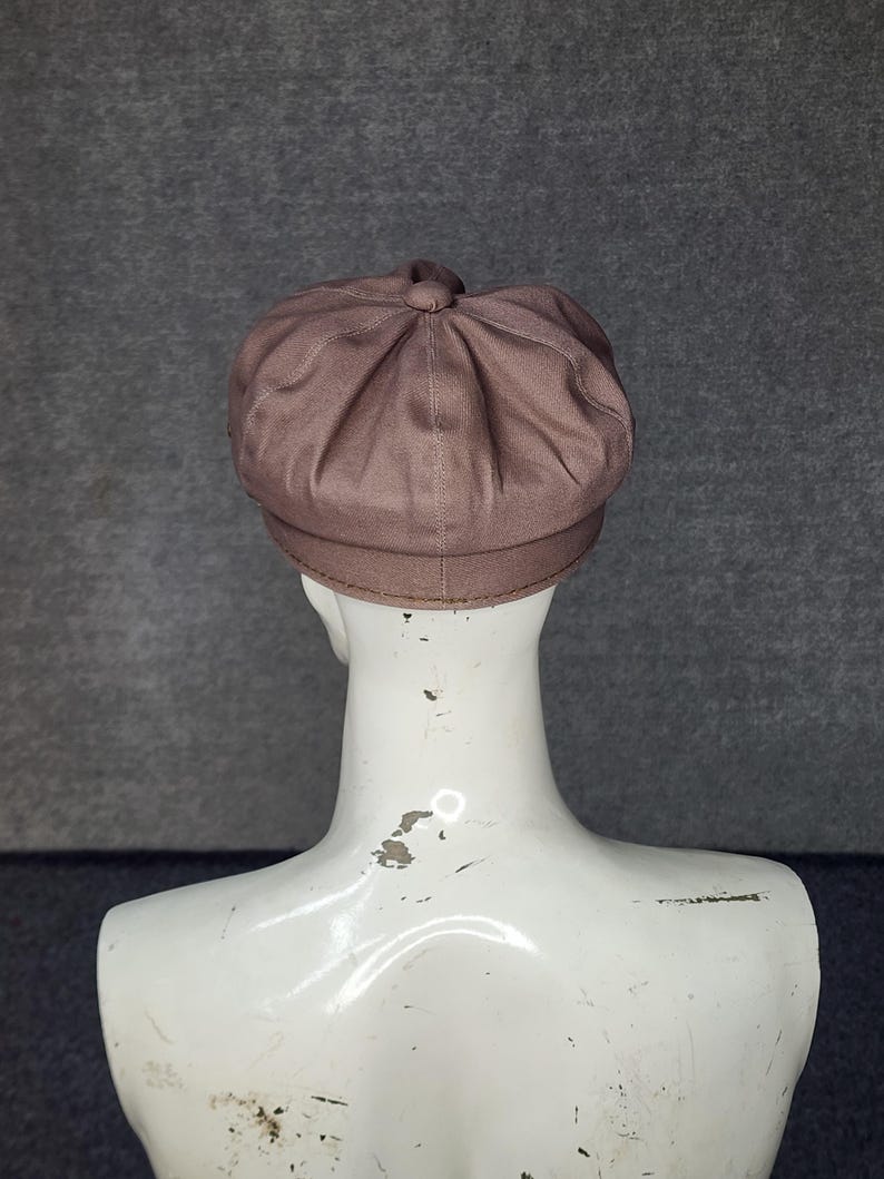 May include: A brown newsboy cap with a rounded crown and a small brim. The cap is made of a soft, textured fabric and has a button detail at the top. The cap is displayed on a white mannequin head against a gray background.