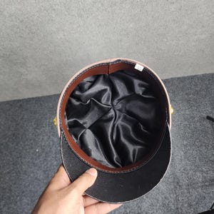 May include: A tan and black military-style hat with a black brim and a brown leather band. The interior is lined with black satin. The hat has gold-colored accents on the sides. The hat is being held by a hand.